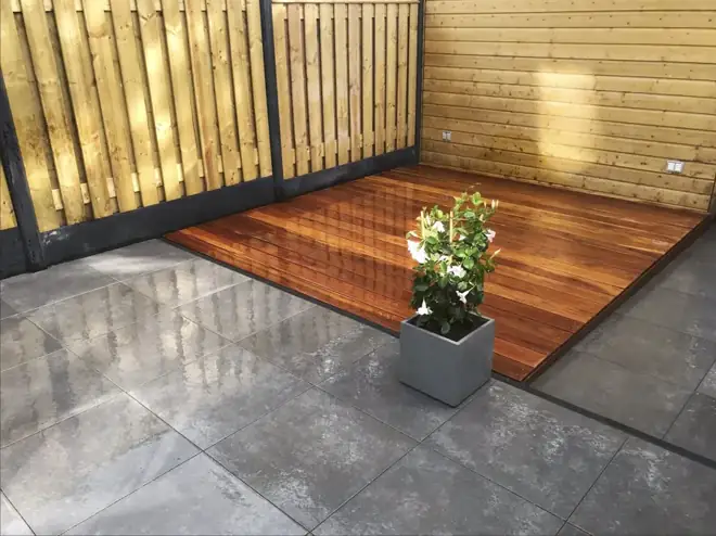 Een houten vlonder naast strakke bestrating, omringd door een houten erfafscheiding. Warme materialen voor een beschutte plek buiten.