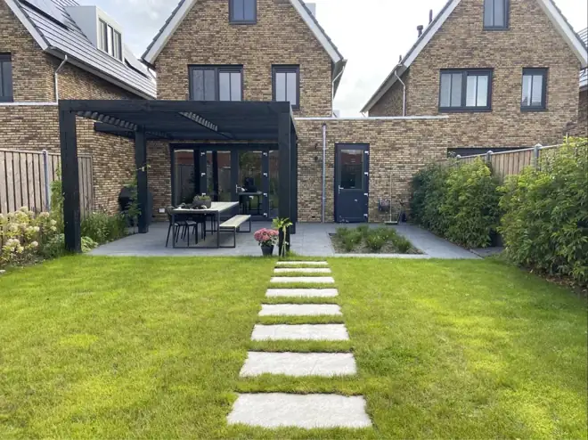 Een groen gazon met een pad van grote tegels naar het terras en de overkapping. Beplanting langs de randen maakt de tuin compleet.