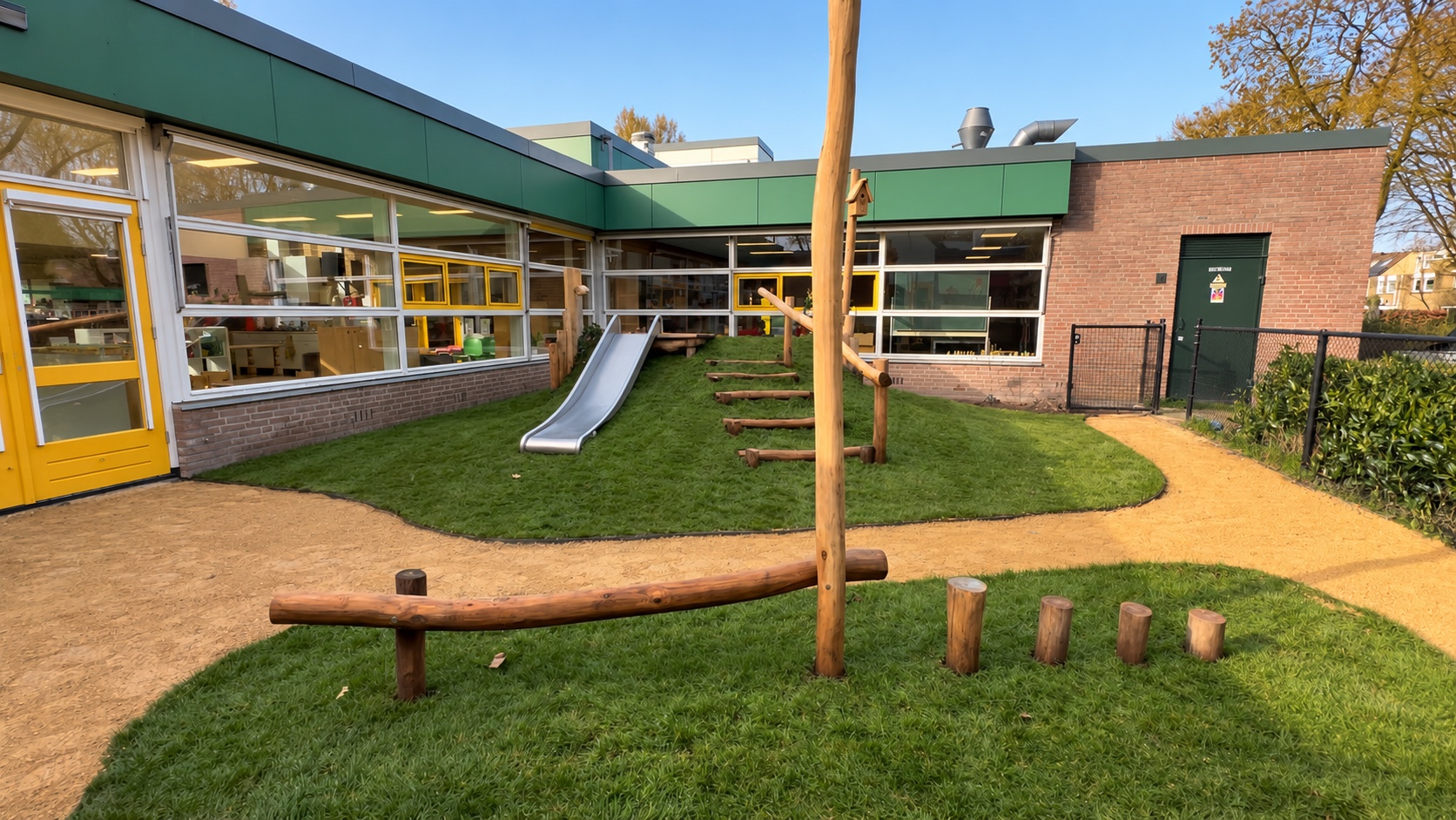 Een groene speelplek met gras, houten klim- en balanceerelementen en een glijbaan. Slingerende paden verbinden de verschillende speelplekken.