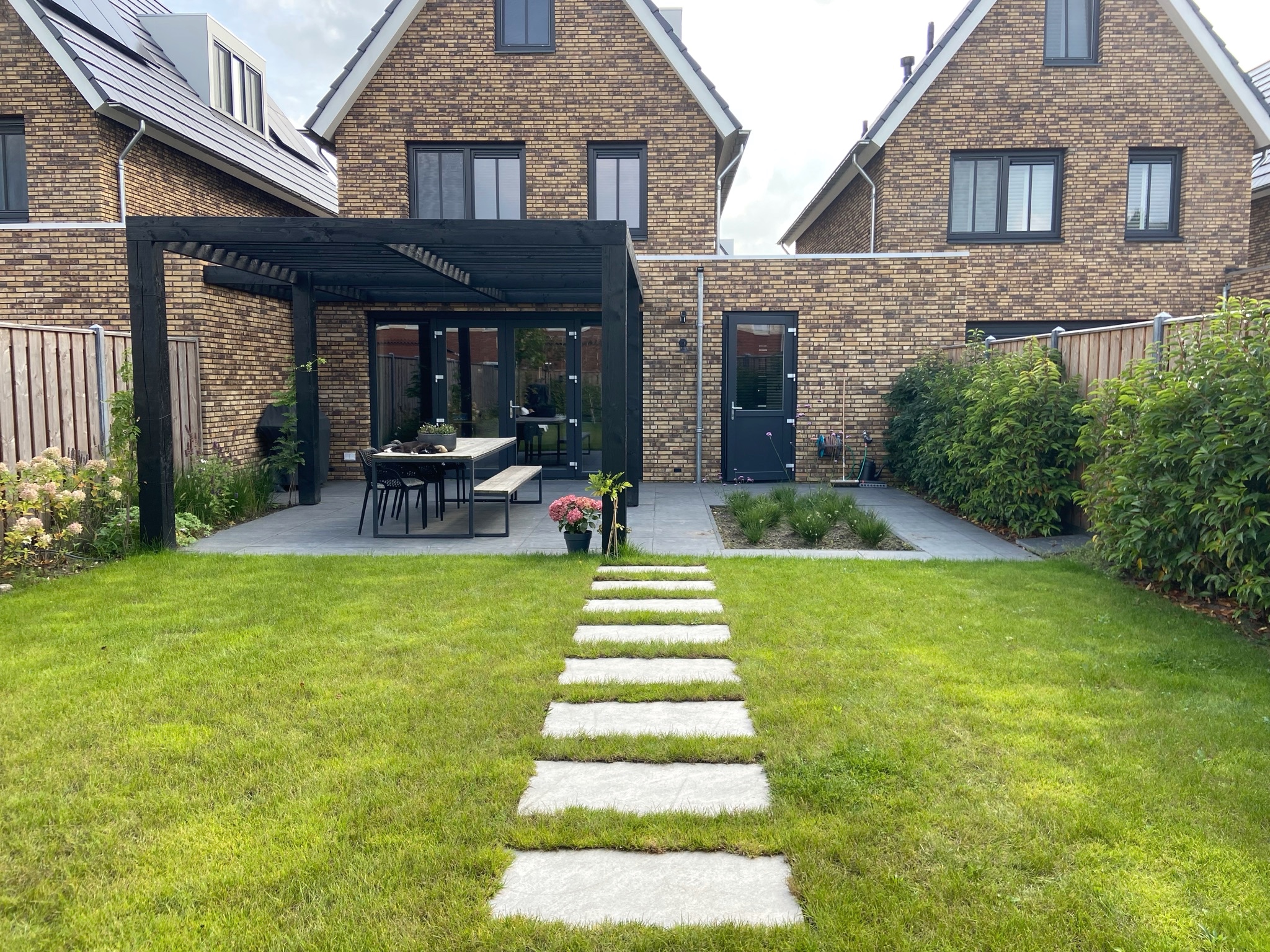 Een groen gazon met een pad van grote tegels naar het terras en de overkapping. Beplanting langs de randen maakt de tuin compleet.